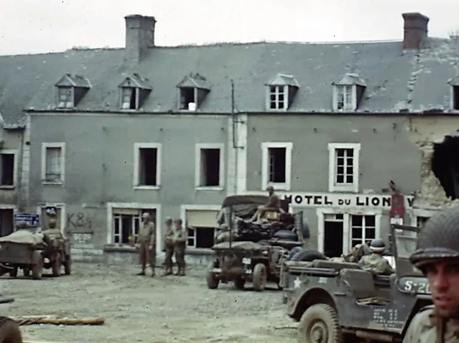 U.S. troops drive through a town by a sign for the Hotel du Lion during World War II in France.webp